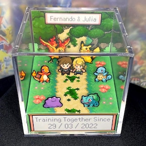Può includere: Una scatola espositiva in acrilico trasparente con un diorama in pixel art di personaggi dei Pokemon e due figure. L'etichetta superiore recita "Fernando & Julia", e l'etichetta inferiore recita "Training Together Since 29/03/2022."