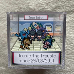 Puede incluir: Una vitrina de acrílico transparente con un diorama pixelado. El texto dice "Team Smith" y "Double the Trouble since 29/08/2011." La escena presenta personajes de dibujos animados y una estética de videojuego retro.