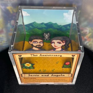 May include: A clear acrylic box featuring a diorama scene with pixelated characters and a landscape. The box is labeled "The Anniversary" with the names "Janie and Angela" below. The scene includes a couple, a cat, and a mountain backdrop.