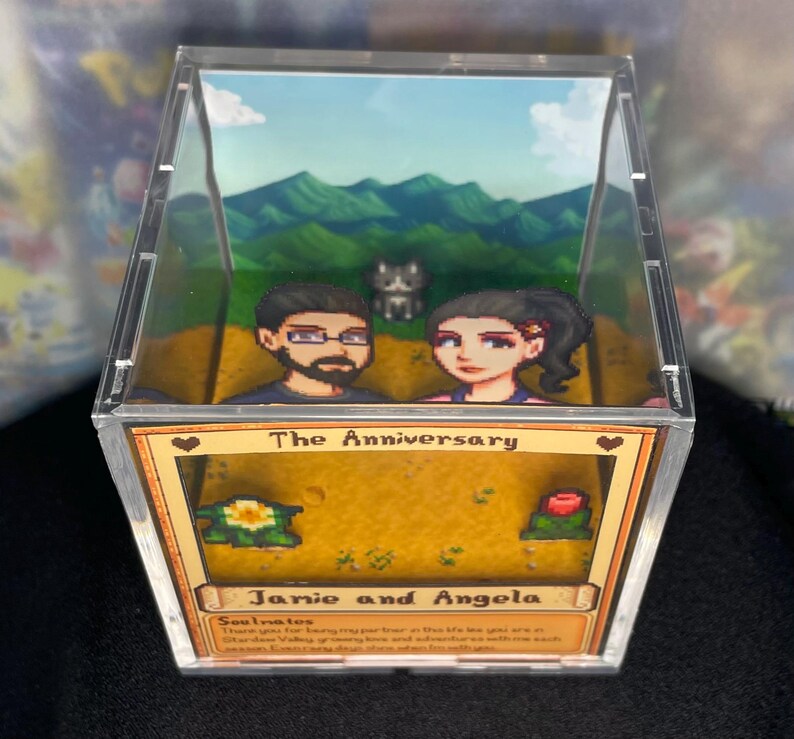 May include: A clear acrylic box featuring a diorama scene with pixelated characters and a landscape. The box is labeled "The Anniversary" with the names "Janie and Angela" below. The scene includes a couple, a cat, and a mountain backdrop.