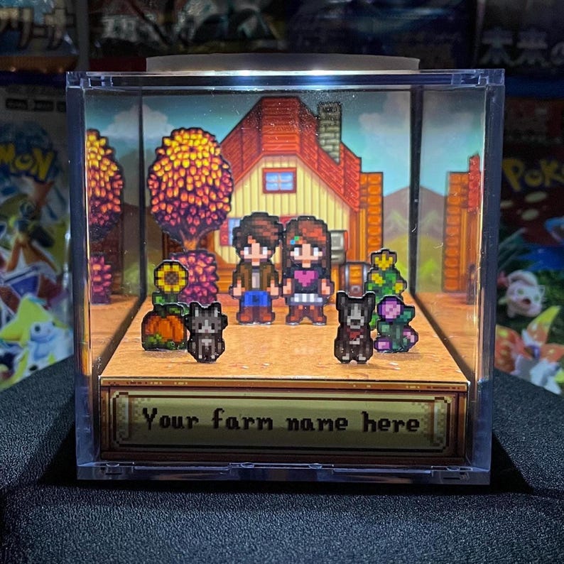 May include: A pixel art diorama in a clear acrylic box featuring a farm scene with a house, trees, and characters. The base has the text "Your farm name here". Includes two pixelated dogs, a pumpkin, and flowers.