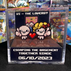 Könnte beinhalten: Ein Pixel-Art-Diorama mit zwei Figuren und einem roten Herzen, in einer transparenten Vitrine. Der Text "VI - THE LOVERS?" steht über den Figuren. Auf der Basis steht "ESCAPING THE BASEMENT TOGETHER SINCE 06/10/2023".