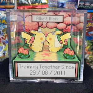 Op de afbeelding: Een heldere acrylbox met een huwelijksscène met twee gele Pikachu-personages die kussen. De scène bevat een pad, roze bloemen en de tekst "Alba & Aleix" en "Training Together Since 29/08/2011".