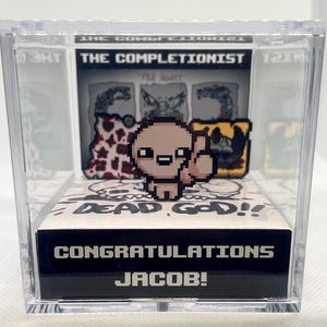 Op de afbeelding: Een transparante acryl vitrine met een zwart-wit 3D-diorama van het videospel "The Binding of Isaac: Rebirth". Het diorama toont het hoofdpersonage, Isaac, met een middelvinger omhoog, en de tekst "Dead God!" De vitrine heeft een zwarte basis met de tekst "Congratulations Jacob!"