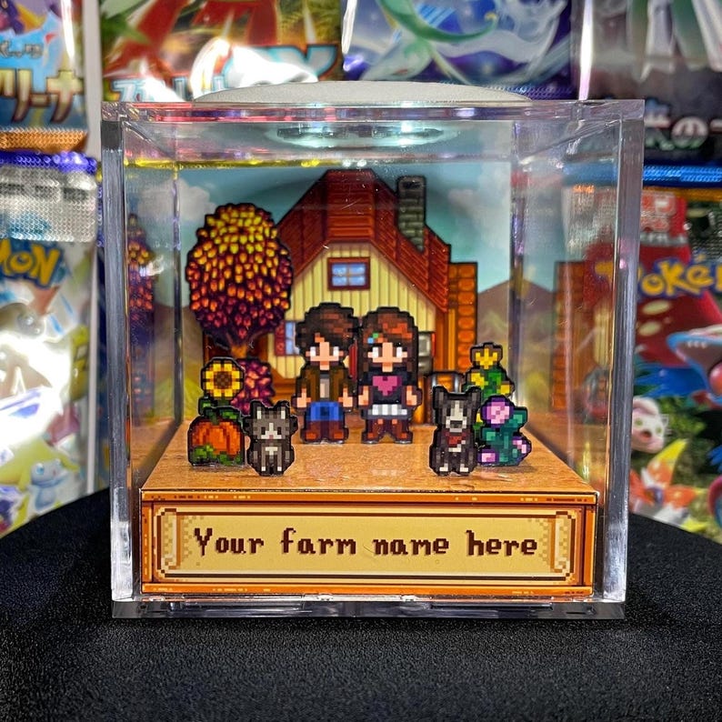 May include: A pixelated diorama inside a clear acrylic cube. The scene depicts a farm with a house, two characters, two dogs, and crops. The text "Your farm name here" is displayed on a gold-colored plaque.