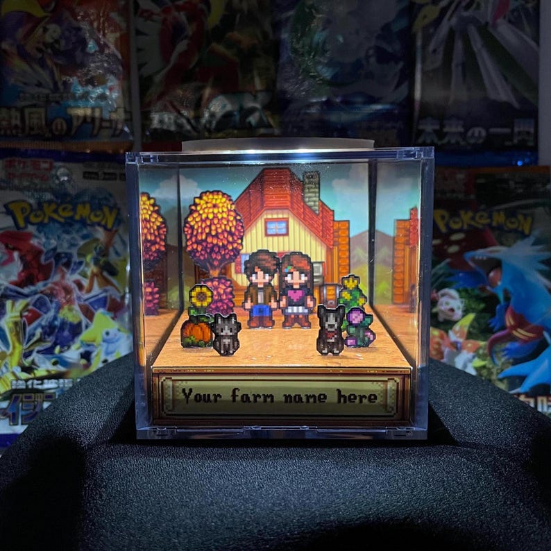 May include: A pixel art diorama in a clear box featuring a house, two characters, two dogs, and various crops. The text "Your farm name here" is displayed on a banner at the bottom. The background includes Pokemon cards.