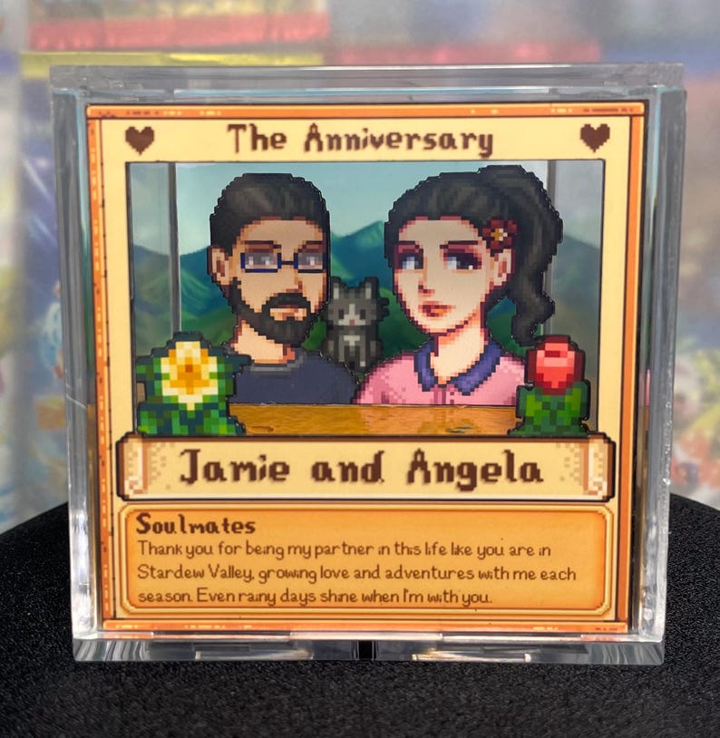May include: A framed anniversary card featuring pixelated artwork of a couple and a cat. The card reads "The Anniversary" and "Jamie and Angela." Text below says "Soulmates" and a message about love and adventures.