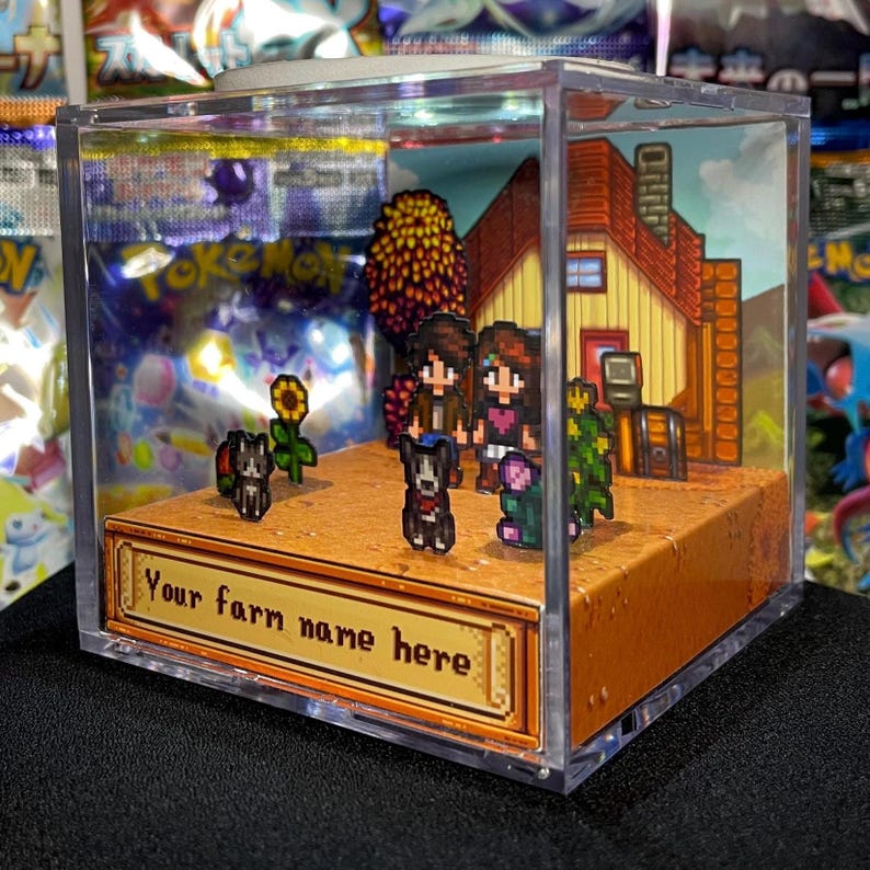 May include: A clear acrylic display case featuring a diorama of a pixelated farm scene. The scene includes a house, characters, dogs, and a sunflower. The base of the display reads "Your farm name here".