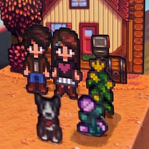 May include: Pixelated figurines of characters and objects from a video game are arranged on a brown surface. The figurines include two people, a dog, a flower, and a robot-like figure. A pixelated house and tree are in the background.