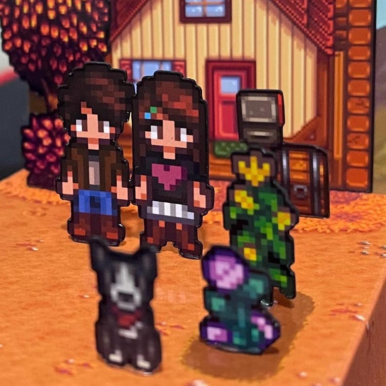 May include: Pixelated figurines of characters and objects from a video game are arranged on a brown surface. The figurines include two people, a dog, a flower, and a robot-like figure. A pixelated house and tree are in the background.