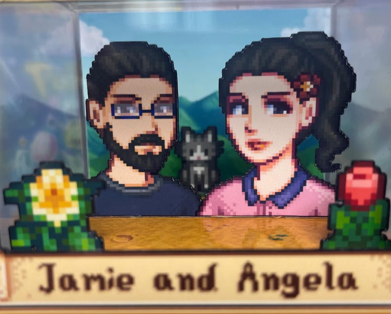 May include: A pixelated diorama featuring two figures, a man with glasses and a beard, and a woman with a flower in her hair. A cat is between them. The base reads "Jamie and Angela". The background shows a mountain scene.