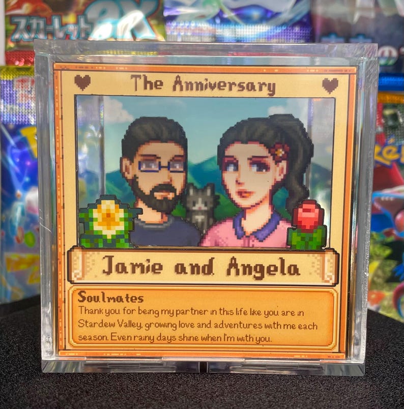 May include: A framed, pixelated art piece featuring a couple and a cat, with the text "The Anniversary" at the top. The names "Jamie and Angela" are displayed below the couple. The bottom text reads "Soulmates" and a loving message.