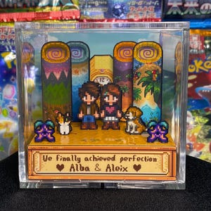 Könnte beinhalten: Ein Pixel-Art-Diorama in einer transparenten Acrylbox. Die Szene zeigt zwei Figuren, zwei Haustiere und eine Landschaft mit dem Text "We finally achieved perfection Alba & Aleix". Der Hintergrund umfasst stilisierte Bäume und eine Uhr.