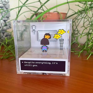 Puede incluir: Un cubo de plástico transparente con un diorama en su interior que representa a un personaje pixelado de un videojuego de pie en una habitación con flores amarillas. El texto "* A pesar de todo, eres tú." está impreso sobre un fondo negro en la parte inferior del cubo.