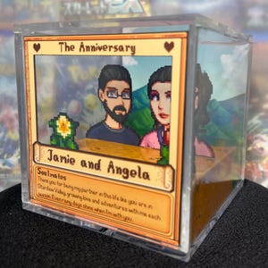May include: A clear acrylic box featuring a pixelated image of a couple, with the text "The Anniversary" and "Jamie and Angela." The box contains a 3D diorama with a Stardew Valley theme, including a flower and a mountain backdrop.