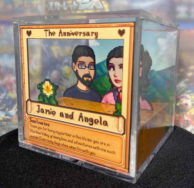 May include: A clear acrylic box featuring a pixelated image of a couple, with the text "The Anniversary" and "Jamie and Angela." The box contains a 3D diorama with a Stardew Valley theme, including a flower and a mountain backdrop.