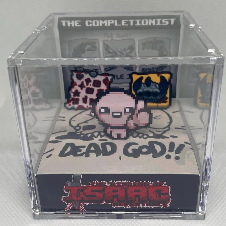 Op de afbeelding: Een transparante acryl kubus display case met een 3D-diorama van het videospel The Binding of Isaac: Rebirth. Het diorama toont een roze personage met een zwarte schedel en de tekst "Dead God!" De onderkant van de case heeft het logo van het spel.