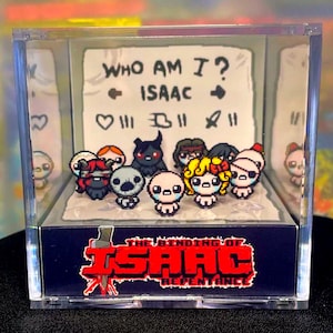 Peut inclure: Un présentoir en acrylique transparent avec des figurines pixélisées de "The Binding of Isaac: Repentance". Le présentoir comprend une toile de fond en papier avec le texte "WHO AM I? ISAAC" et divers symboles. La base porte le logo du jeu en rouge et blanc.