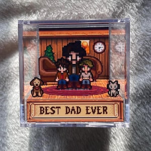 Könnte beinhalten: Transparente Acryl-Displaybox mit einer pixeligen Familienszene: ein Vater und zwei Kinder, ein Hund und eine Katze. Der Text "BEST DAD EVER" ist auf der Vorderseite zu sehen. Die Szene spielt in einem gemütlichen Raum.