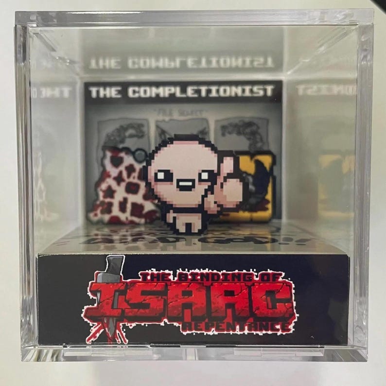 Op de afbeelding: Een transparante acryl vitrine met een klein beeldje van Isaac uit het videospel The Binding of Isaac: Repentance. Het beeldje is roze en heeft een gepixelde vormgeving. De vitrine bevat ook een klein kaartje met de tekst "The Completionist" en een klein kaartje met de tekst "The Binding of Isaac: Repentance".