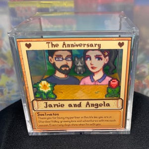 May include: A clear acrylic box containing a framed image with the text "The Anniversary" and the names "Jamie and Angela." The image features pixelated artwork of a couple and a cat, with a message below.