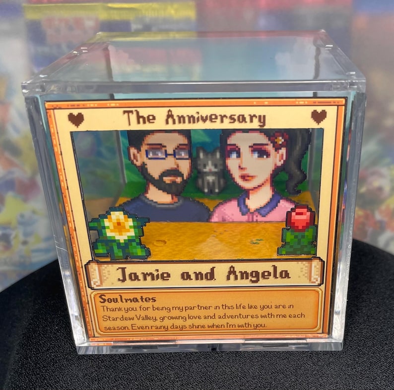 May include: A clear acrylic box containing a framed image with the text "The Anniversary" and the names "Jamie and Angela." The image features pixelated artwork of a couple and a cat, with a message below.