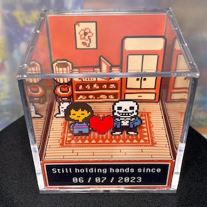 Può includere: Diorama in arte pixelata con personaggi di Undertale, all'interno di una scatola di acrilico trasparente. La scena mostra Frisk e Sans con un cuore rosso, con il testo "Still holding hands since 06/07/2023" sulla base.