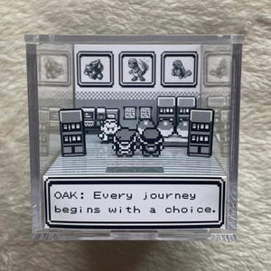 Op de afbeelding: Een heldere acryl display kubus met een gepixeld tafereel uit een videogame. De scène bevat personages, een tekstvak met de quote "OAK: Elke reis begint met een keuze" en afbeeldingen van drie wezens.