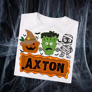May include: White t-shirt featuring a Halloween-themed design. The design includes a pumpkin wearing a witch hat, Frankenstein's monster, and a mummy. The name "AXTON" is displayed in a brown frame with a spider.