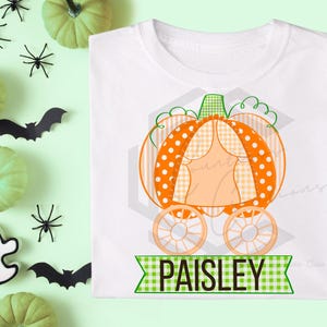 Puede incluir: Camiseta blanca con un diseño de carroza de calabaza, con lunares naranjas y patrones de cuadros. El nombre "PAISLEY" se muestra en una pancarta verde. Decoraciones de Halloween, incluyendo calabazas, murciélagos, arañas y un fantasma, rodean la camiseta.