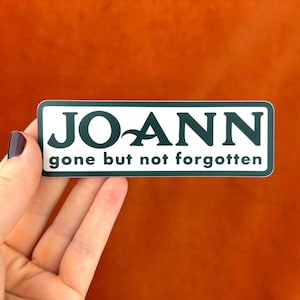 Puede incluir: Una pegatina rectangular con fondo blanco y borde verde oscuro. La pegatina dice "JOANN" en letras negritas verde oscuro, con un gráfico de onda estilizado. Debajo, el texto "gone but not forgotten" está impreso en una fuente más pequeña.