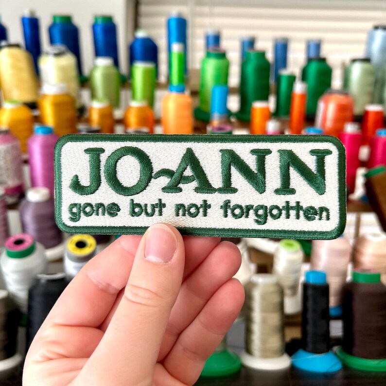 May include: A rectangular embroidered patch with the name "JO-ANN" in green, with the text "gone but not forgotten" below. The patch is white with a green border. The background features spools of colorful thread.
