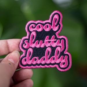 May include: A pink embroidered patch with the words "cool slutty daddy" in a cursive font. The patch has a black background and a pink border. The patch is held up against a blurred green background.
