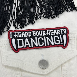 Puede incluir: Chaqueta vaquera blanca con flecos negros en la parte superior. Un parche en la chaqueta dice "I HEARD YOUR HEARTS DANCING!" en texto blanco con borde rojo.