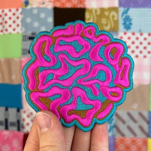 Puede incluir: Parche bordado rosa y turquesa con la palabra "Brain" en una fuente ondulada y abstracta.