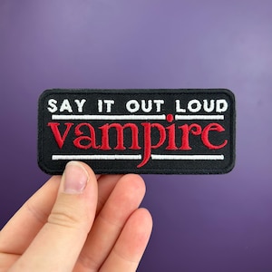 Puede incluir: Parche rectangular negro con las palabras "SAY IT OUT LOUD" en blanco sobre la palabra "vampire" en rojo. El parche tiene un borde blanco. Fondo morado.
