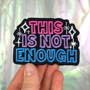 Peut inclure: Écusson brodé avec le texte "THIS IS NOT ENOUGH" en rose, violet et bleu. L'écusson a une bordure noire avec des accents d'éclats d'étoiles et de diamants blancs. L'écusson est tenu par une main.