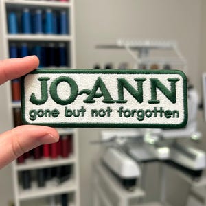 May include: Embroidered patch with the name "JO-ANN" in green, with the phrase "gone but not forgotten" below. The patch has a white background and a green border. The patch is held in front of a blurred background.