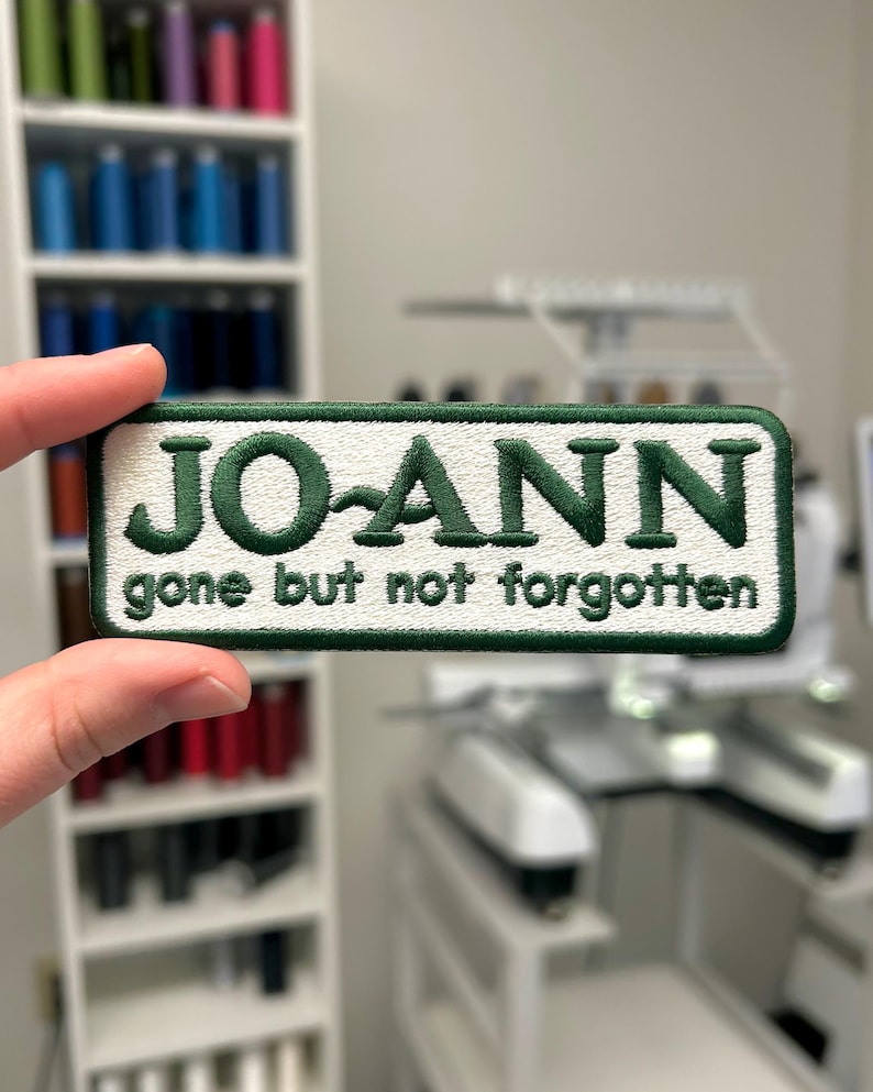 May include: Embroidered patch with the name "JO-ANN" in green, with the phrase "gone but not forgotten" below. The patch has a white background and a green border. The patch is held in front of a blurred background.