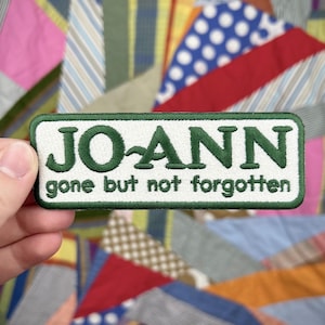 May include: A rectangular patch with the name "JO-ANN" embroidered in green on a white background. The text "gone but not forgotten" is below the name. The patch has a green border and is held against a colorful patchwork background.