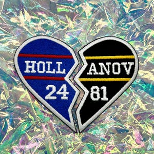 Op de afbeelding: Een gebroken hart patch met blauwe en zwarte helften. De blauwe helft leest "HOLL 24" met rode en witte accenten. De zwarte helft leest "ANOV 81" met gele en witte accenten. De patch ligt op een holografische achtergrond.