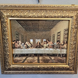 May include: A framed print of The Last Supper featuring a central figure seated at a long table with twelve individuals in a room with a coffered ceiling. The artwork is enclosed in an ornate, rectangular gold-colored frame with relief detailing.