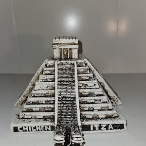 May include: A white and brown decorative replica of the Chichen Itza pyramid. The pyramid has a detailed staircase and a small temple at the top. The words "CHICHEN ITZA" are printed on the base.
