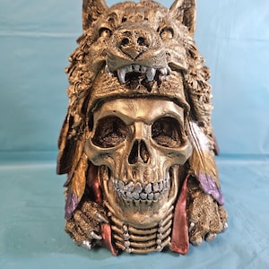 May include: A gold-colored skull sculpture with a wolf headdress. The skull has silver teeth and is adorned with feathers in shades of purple and red. The wolf headdress is detailed with textured fur and sharp teeth.
