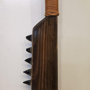 May include: A wooden club with a dark brown stained finish featuring a row of sharp, black obsidian-like blades embedded along one side. The handle is wrapped in a tan leather cord, providing a textured grip for the long, straight weapon.