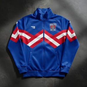 Puede incluir: Chaqueta deportiva azul con diseño de chevrón rojo y blanco en el pecho. Cuenta con cremallera completa, dos bolsillos delanteros y un pequeño emblema con la palabra "HAITI" en el pecho superior. Un logotipo blanco está en el pecho izquierdo.
