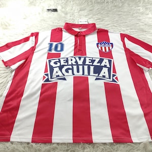 Op de afbeelding: Rood en wit gestreept voetbalshirt met het nummer 10 in blauw. Het shirt heeft de tekst "CERVEZA AGUILA" en een teamembleem. De kraag en mouwen zijn rood en wit gestreept.