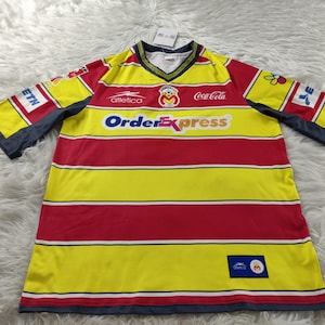 May include: Yellow and red striped soccer jersey with the words "OrderExpress" and "Coca-Cola" on the front. The jersey has a blue logo with a cartoon bear on the left side of the chest.