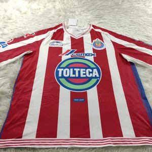 Może przedstawiać: Piłkarska koszulka w czerwono-białe pasy z logo drużyny i sponsorami, "Atletica", "Cemex" i "Tolteca" nadrukowanymi z przodu. Koszulka ma niebieskie wykończenie wok&oacute;ł rękaw&oacute;w i dołu.