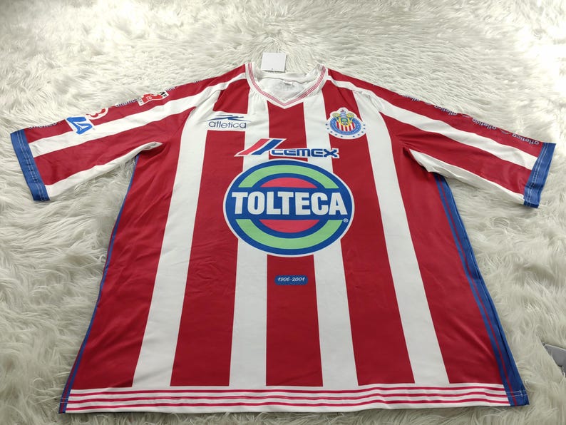 Może przedstawiać: Piłkarska koszulka w czerwono-białe pasy z logo drużyny i sponsorami, "Atletica", "Cemex" i "Tolteca" nadrukowanymi z przodu. Koszulka ma niebieskie wykończenie wok&oacute;ł rękaw&oacute;w i dołu.